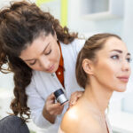 Dermatologist in latex gloves holding dermatoscope while examining attractive patient with skin disease. Female dermatologist examining patient with dermascope, looking for signs of skin cancer.